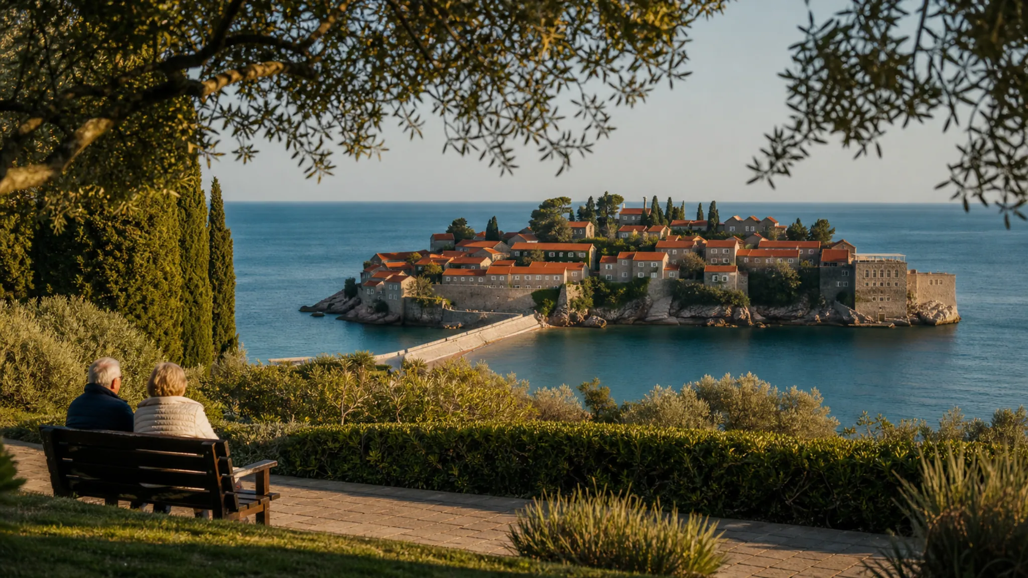 Blick aus dem Miločer-Park auf die Inselfestung Sveti Stefan; ein älteres Paar sitzt auf einer Bank.